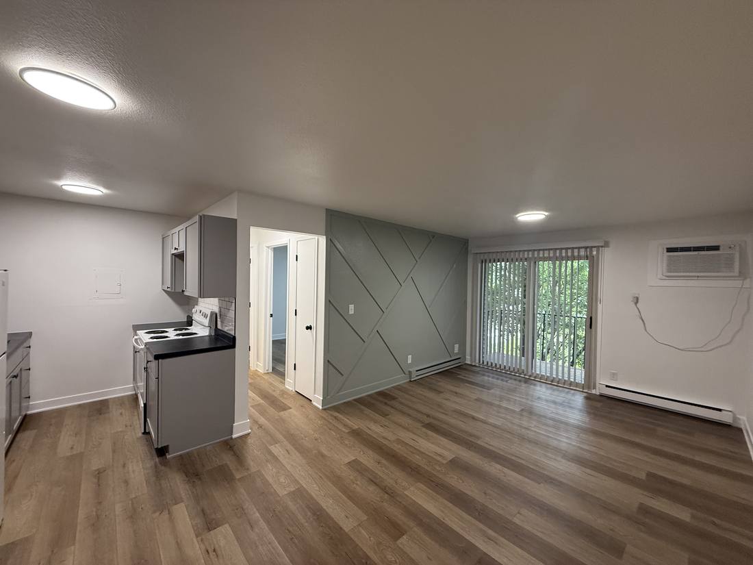 A renovated living room with wide-plank flooring, a painted accent wall in a diagonal batten pattern, and the open kitchen visible to the left.