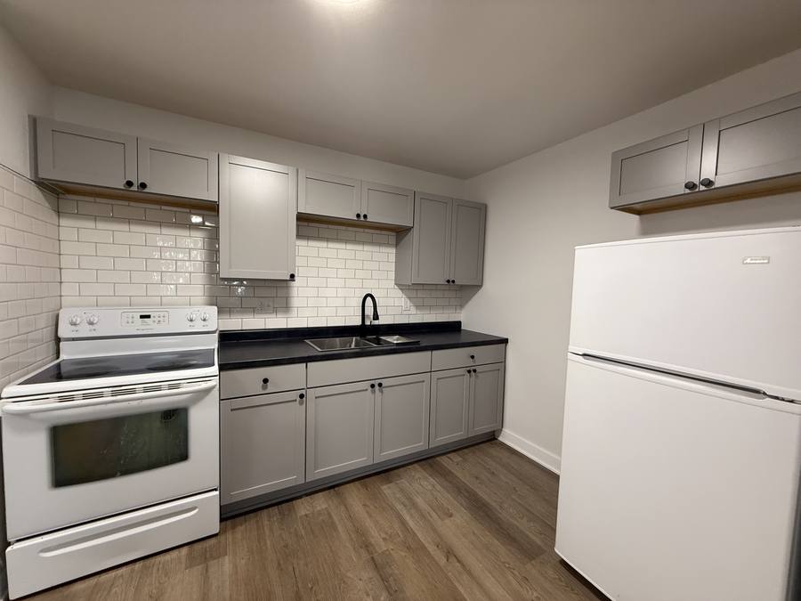 A renovated kitchen with light shaker cabinets, a dark countertop, white subway tile backsplash, an open wood shelf, and a white range and refrigerator.