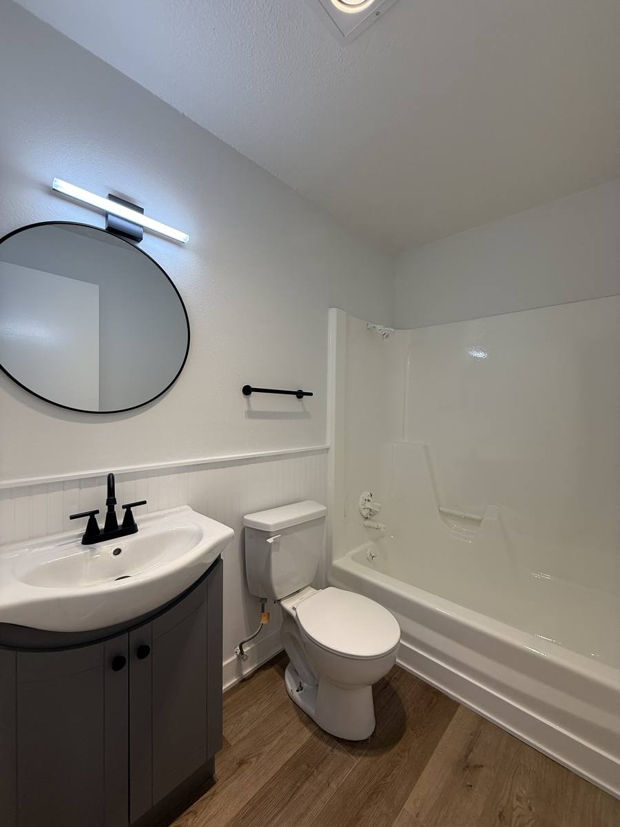 A renovated bathroom with a round black-framed mirror, a black linear light bar, a grey vanity and a white tub and shower surround.