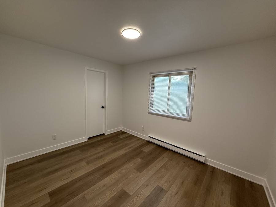 An empty bedroom with wide-plank flooring, a window, and electric baseboard heat along the wall.