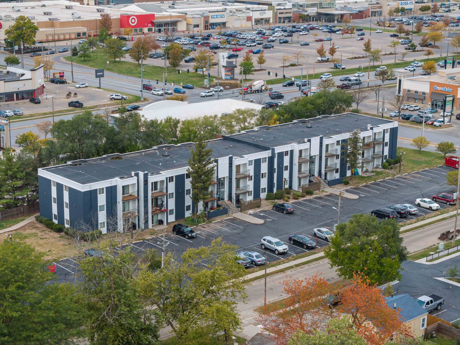 Aerial view of Crossroad Apartments, a long three-story building painted white and slate grey, with the Merle Hay retail corridor and its parking lots directly behind it.